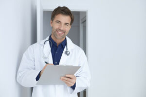 Smiling male doctor in a white coat holding a clipboard, standing in a bright medical office.
