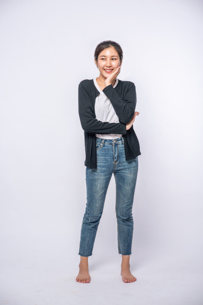 "Smiling young woman in a black cardigan and jeans standing confidently against a white background, radiating happiness and positivity."