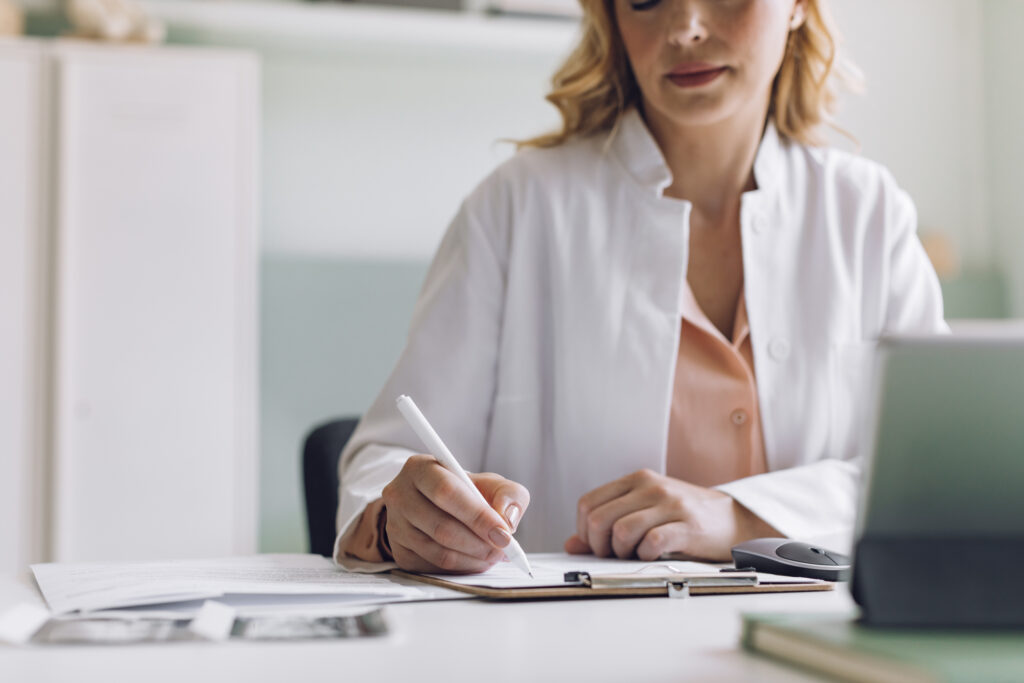 A person in a white coat writes on a clipboard at a desk.