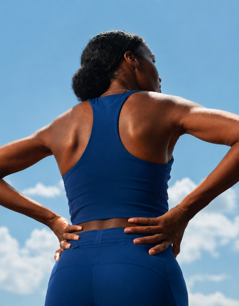 Athletic woman in a blue workout outfit standing outdoors with hands on hips under a bright sky, showcasing strength, confidence, and fitness.
