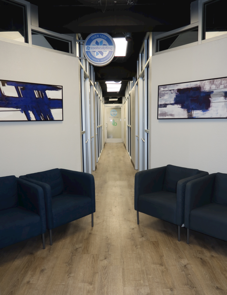 Modern Weight Loss MD clinic hallway with blue lounge chairs, contemporary artwork, wood flooring, and private treatment rooms leading down a well-lit corridor.