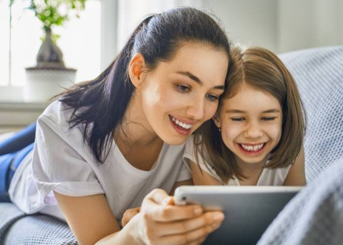 A smiling woman and a young girl lie on a couch together, happily looking at a tablet screen. They appear to be enjoying what they’re watching as they relax in a bright, cozy living room.