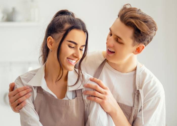 A young man stands behind a smiling woman, gently holding her shoulders as they both wear light aprons in a bright kitchen.