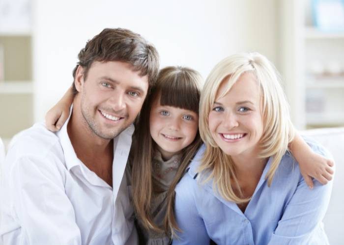 A smiling man and woman sit close together with a young girl between them, her arms around both adults as they pose happily for the camera.